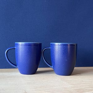 Peut inclure: Deux tasses en céramique bleu cobalt avec une texture mouchetée. Les tasses ont une forme arrondie et une anse incurvée. Elles sont posées sur un fond bleu foncé, sur une surface en bois clair.