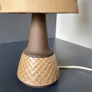 May include: A brown ceramic table lamp with a beige lampshade. The lampshade has a brown floral pattern. The base of the lamp has a textured, diamond-shaped pattern.