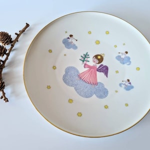 May include: White ceramic plate with a gold rim featuring a festive design of angels and stars. The angels are depicted in pink, blue, and yellow, with one angel holding a sprig of greenery.
