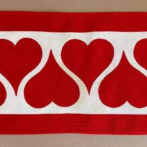 May include: Red and white fabric with a repeating pattern of white hearts on a red background. The fabric has a white border.