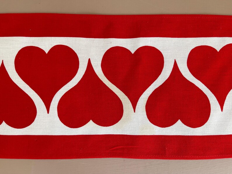 May include: Red and white fabric with a repeating pattern of white hearts on a red background. The fabric has a white border.