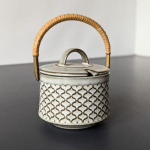 Puede incluir: Un azucarero de cerámica blanca con un patrón geométrico marrón y un asa trenzada. La tapa tiene un pequeño pico.