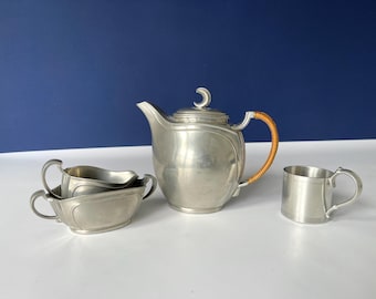 May include: A vintage pewter tea set, including a teapot with a woven handle, a creamer, a sugar bowl, and a small mug. The set is displayed on a white surface against a dark blue background. The teapot has a crescent moon finial.