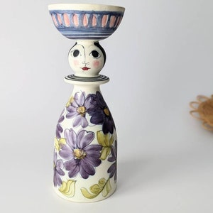 Könnte beinhalten: Eine Keramikfigur mit einem stilisierten Gesicht und einem violetten Blumenmuster. Die Figur hat eine Schale auf dem Kopf.
