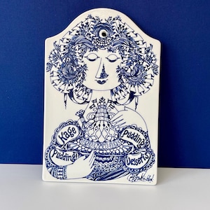 Puede incluir: Placa decorativa de cerámica blanca con una ilustración azul de una mujer sosteniendo un postre. La placa tiene las palabras "Kage Pudding Desserts" en un diseño circular. La cara de la mujer está centrada, con los ojos cerrados y cabello elaborado.
