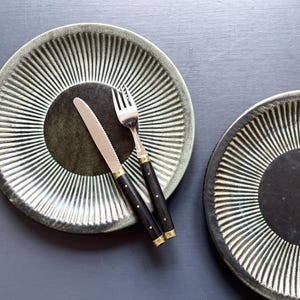 May include: Overhead shot of two ceramic plates with a radial design in white and dark green. A knife and fork with black handles and brass accents rest on one plate. The plates are on a dark gray surface.