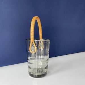 May include: A clear glass ice bucket with a woven handle. The bucket has a gray tint and etched floral designs. The handle is made of woven tan material. The ice bucket is on a white surface with a dark blue background.