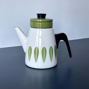 May include: White enamel coffee pot with a green lid and leaf design. The pot has a black handle and spout. A vintage kitchenware item.