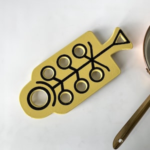 Può includere: Un sottopentola giallo con un design astratto nero e ritagli circolari. Accanto, una casseruola in acciaio inossidabile con bordo in rame e manico in ottone. Il sottopentola serve a proteggere le superfici.