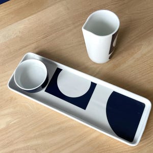 May include: A white ceramic serving tray with a navy blue and white geometric design, accompanied by a small bowl and a creamer. The tray is rectangular, and the creamer has a dark brown accent.
