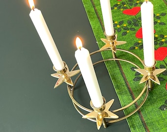 May include: A brass candle holder with four lit white candles. The candle holder has a circular design with star-shaped holders. The candles are on a dark surface with a green runner with red hearts.