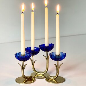 Könnte beinhalten: Ein goldfarbener Kerzenleuchter aus Metall mit vier Kerzenhaltern. Jeder Halter ist in Form einer blauen Blume mit einer weißen Kerze darin. Die Kerzen sind angezündet.
