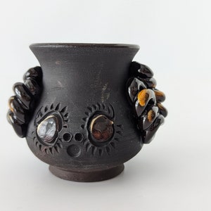 May include: A small, black ceramic pot with two sculpted handles. The pot has a decorative design of stylized eyes and circles.