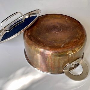 May include: A round copper pot with a polished silver-tone metal handle and a matching lid featuring a silver-tone handle. The pot has a warm, metallic finish with visible signs of use and oxidation, showcasing a classic kitchen cookware design.