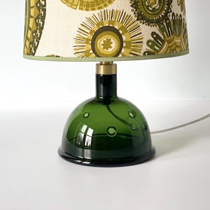 May include: A green glass table lamp with a patterned lampshade. The base is a dark green bottle shape with circular indentations. The lampshade features a cream background with olive green and yellow floral designs. A brass fitting connects the base to the lampshade.