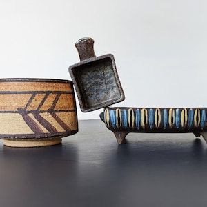 May include: Three ceramic planters with geometric designs. The planter on the left has brown and tan stripes with a geometric pattern. The planter in the middle is brown with a blue and white geometric pattern. The planter on the right is brown with blue and yellow stripes.