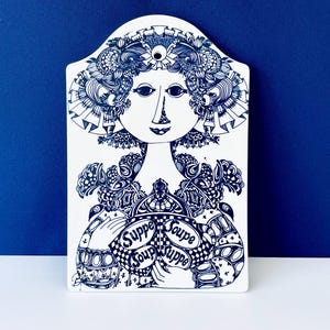 Puede incluir: Placa de pared de cerámica blanca con una ilustración azul y blanca de una mujer con un tocado y ropa floral ornamentada. Las palabras "Suppe Soup" están en un círculo. La placa tiene una parte superior curva.