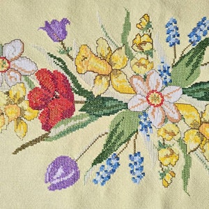 May include: A cross-stitch floral pattern showcasing a vibrant array of flowers. The design includes daffodils, tulips, and other blooms in yellow, red, purple, and white, set against a pale yellow backdrop.