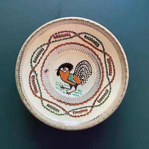 May include: A round, ceramic plate with a rooster design. The plate features a white base with a central rooster illustration in black, orange, and green. The rim is decorated with a repeating pattern of brown, green, and red. The plate is set against a dark teal background.
