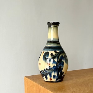 May include: A ceramic vase with a cream-colored base, adorned with blue floral patterns and accents. The vase has a dark blue rim and a slender neck. Small brown dots are scattered across the design. The vase is displayed on a wooden surface.