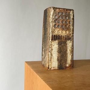 Puede incluir: Un jarrón rectangular de cerámica texturizada en tonos marrón, crema y beige. El jarrón presenta hendiduras circulares y líneas verticales, lo que le da un aspecto artesanal. Se encuentra sobre una superficie de madera. El jarrón mide unos 20 cm de alto.