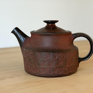 May include: A dark reddish-brown ceramic teapot with a glossy black spout and handle. The body features a repeating embossed pattern of stylized faces in square grids. It includes a matching lid with a small knob handle on top.