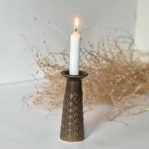 Puede incluir: Una vela blanca encendida en un candelabro de color bronce. El candelabro tiene un patrón floral texturizado. En el fondo hay follaje seco de color marrón claro. La llama de la vela es brillante.