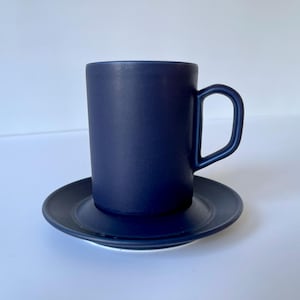 Puede incluir: Un juego de taza y platillo de cerámica azul oscuro. La taza tiene un diseño sencillo y un asa redondeada. El platillo es ligeramente más grande que la taza y tiene una superficie lisa.
