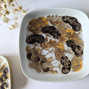 May include: A square ceramic dish with a white background and a brown and yellow floral pattern. The dish has a slightly raised rim.