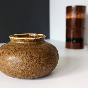 Könnte beinhalten: Eine kleine, runde, braune Keramikvase mit einem gesprenkelten Überzug. Die Vase hat einen weißen Rand und steht auf einer weißen Oberfläche.