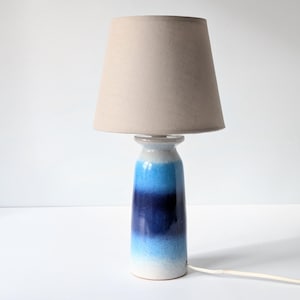 May include: A blue and white ceramic table lamp with a beige lampshade. The lamp base has a gradient of blue shades, from light blue to dark blue.
