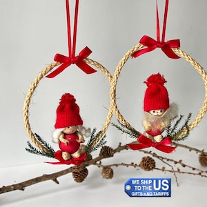 Op de afbeelding: Twee kerstversieringen met kleine kabouterfiguren met rode mutsen en outfits, geplaatst in geweven touw cirkels. Elk ornament is versierd met een rode strik en wordt opgehangen door een rood lint. De ornamenten worden gepresenteerd op een tak met dennenappels.
