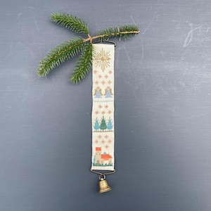 Könnte beinhalten: Handgestickter Weihnachtsschmuck mit cremefarbenem Hintergrund, mit einem goldenen Stern, Engeln, Bäumen und einer Kirche. Am unteren Ende hängt eine kleine Messingglocke. Oben befindet sich ein Tannenzweig. Der Schmuck ist ca. 25 cm lang.