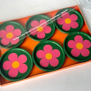 Op de afbeelding: Set van zes ronde onderzetters met een retro bloemenontwerp. Elke onderzetter heeft een roze bloem met een gele kern op een groene achtergrond. De onderzetters zitten in een oranje doos met een transparante plastic verpakking.