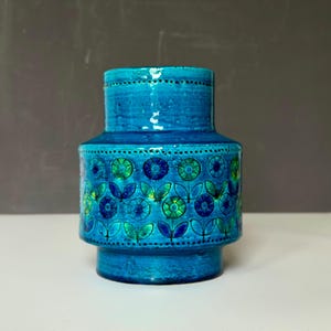Puede incluir: Un jarrón de cerámica turquesa con forma cilíndrica y base ancha. El jarrón presenta una superficie texturizada y está adornado con un estampado floral en tonos azules y verdes. El jarrón mide unos 20 cm de altura.