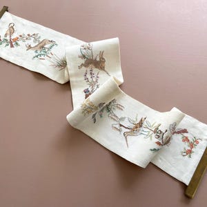 May include: A white linen tapestry with embroidered images of birds and animals, including a rabbit, a bird, and a deer. The tapestry is mounted on a wooden frame with brass hardware.
