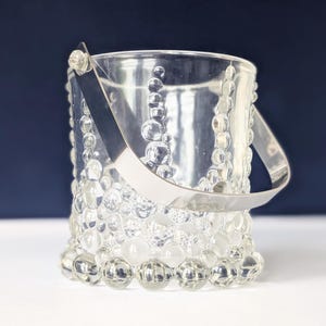 Op de afbeelding: Een heldere glazen ijsemmer met een zilveren handvat. De emmer is versierd met een bubbelpatroon en het handvat is bevestigd met zilveren klinknagels. Ontworpen voor het koelen van dranken.