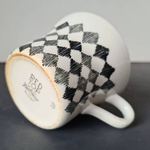 Op de afbeelding: Witte keramische theekop met een zwart-grijs geruit patroon. De onderkant van de kop heeft de tekst "Red Top Pottery England" in zwart gedrukt.