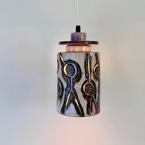 May include: A cylindrical ceramic pendant light fitting featuring a white glazed surface with dark brown, incised abstract stick figure motifs. The top section includes a layered, iridescent purple and copper-toned disc cap with a white cord.