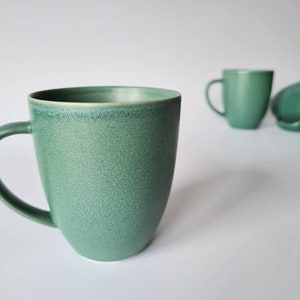 Puede incluir: Tres tazas de cerámica verde con un acabado moteado. Las tazas tienen una forma redondeada y un asa.
