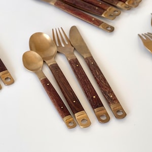 Può includere: Una collezione di posate in stile vintage con teste in metallo color ottone e manici in legno marrone scuro. Il set include cucchiai, forchette e coltelli, ciascuno con un terminale in ottone con un foro. Le posate sono disposte su una superficie bianca.