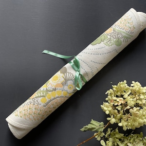 Könnte beinhalten: Ein weißer Tischläufer mit einem floralen Muster in Gelb-, Grün- und Brauntönen. Der Läufer ist aufgerollt und mit einem grünen Band gebunden.