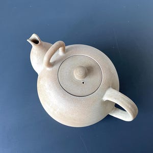 Op de afbeelding: Een beige keramische theepot met een afgeronde body, een gebogen handvat en een deksel met een kleine knop. De tuit is gebogen en omhoog gericht. De theepot staat op een donkerblauwe ondergrond.