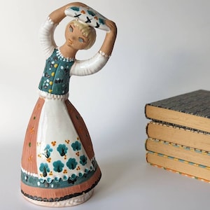 Könnte beinhalten: Eine Keramikfigur einer Frau in einem grün-weiß-rosa Kleid mit Blumenmustern. Sie hält einen weißen Teller über ihrem Kopf und befindet sich in einer Tanzpose.