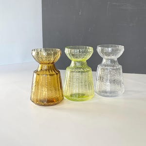 Op de afbeelding: Drie glazen vazen in amber, limoengroen en helder. Elke vaas heeft een uitlopende bovenkant en een brede basis, met een textuur, geweven patroon op het lichaam. De vazen zijn gerangschikt op een wit oppervlak tegen een grijze en witte achtergrond.