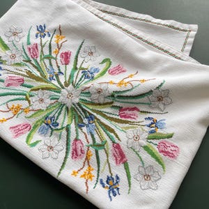 Peut inclure: Nappe en lin blanc avec une broderie florale détaillée. La broderie présente un bouquet de tulipes, de jonquilles et d'iris dans des tons de rose, bleu, jaune et blanc, avec des tiges et des feuilles vertes. Les bords sont finis avec une bordure cousue verte et rouge.