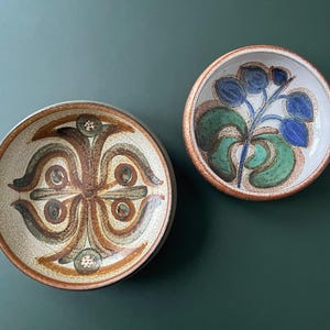 May include: Two ceramic bowls with decorative designs. One bowl features a symmetrical pattern in brown, tan, and green. The other bowl has a floral design in blue and green. Both bowls have a brown rim.
