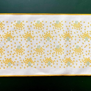 May include: White rectangular table runner featuring a repeating pattern of yellow floral bouquets and scattered flowers. The flowers have green stems and are set against a white background. The runner has a yellow border.