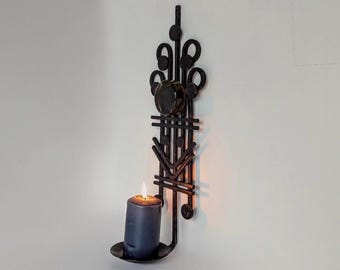 Peut inclure: Un bougeoir mural en métal noir présentant un design géométrique vertical avec des éléments circulaires et linéaires. Une bougie pilier gris foncé est placée dans la coupelle inférieure, avec une flamme allumée projetant une lueur.