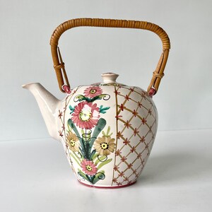 May include: A white ceramic teapot with a floral design and a woven bamboo handle. The teapot has a round body and a spout on the side. The floral design features pink, yellow, and green flowers.